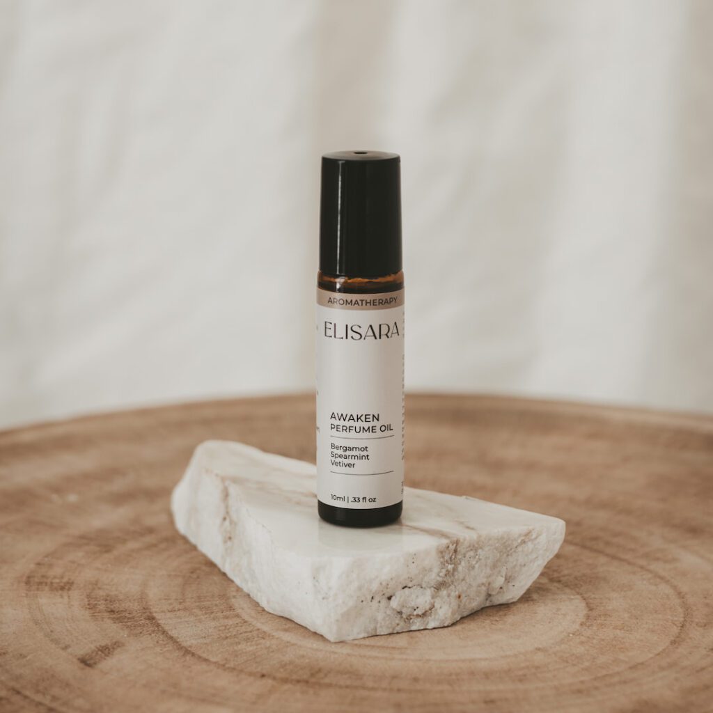 A 10ml amber roller bottle with a black lid and a white label that reads "Awaken perfume oil" with the ingredients bergamot, spearmint, and vetiver. The bottle is placed on a flat, white quartz stone, resting on a round timber stool. In the background, there is a white fabric curtain.