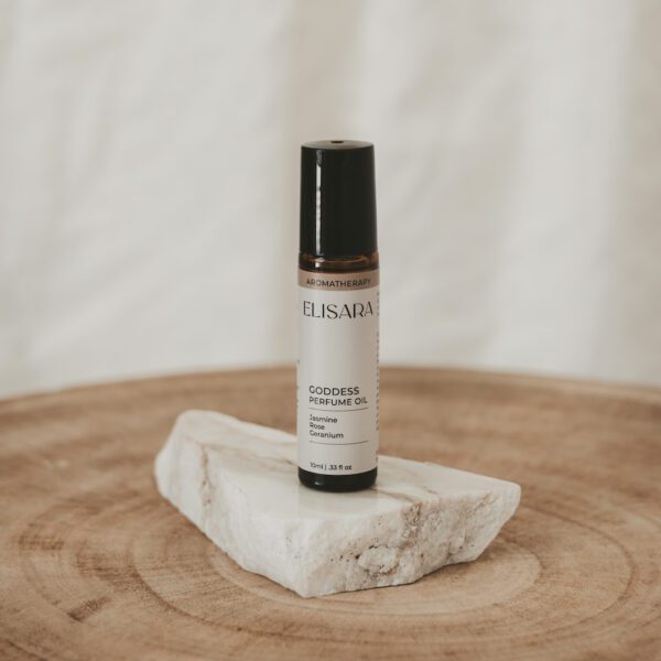 A 10ml amber roller bottle with a black lid and a white label that reads "Goddess perfume oil" with the ingredients Jasmine, Rose and geranium listed. The bottle is placed on a flat, white quartz stone, resting on a round timber stool. In the background, there is a white fabric curtain.