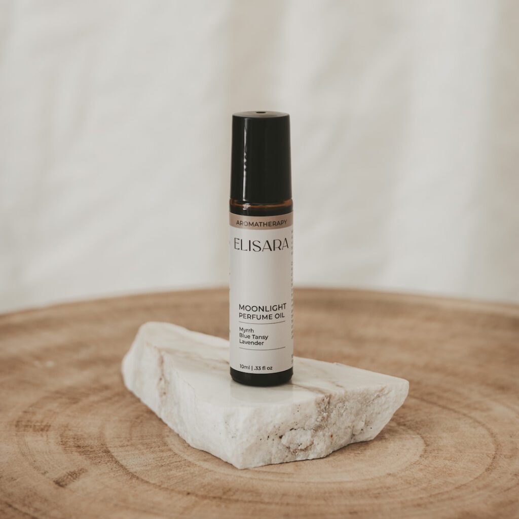 A 10ml amber roller bottle with a black lid and a white label that reads "Moonlight perfume oil" with the ingredients Myrrh, Blue Tansy and lavender listed. The bottle is placed on a flat, white quartz stone, resting on a round timber stool. In the background, there is a white fabric curtain.