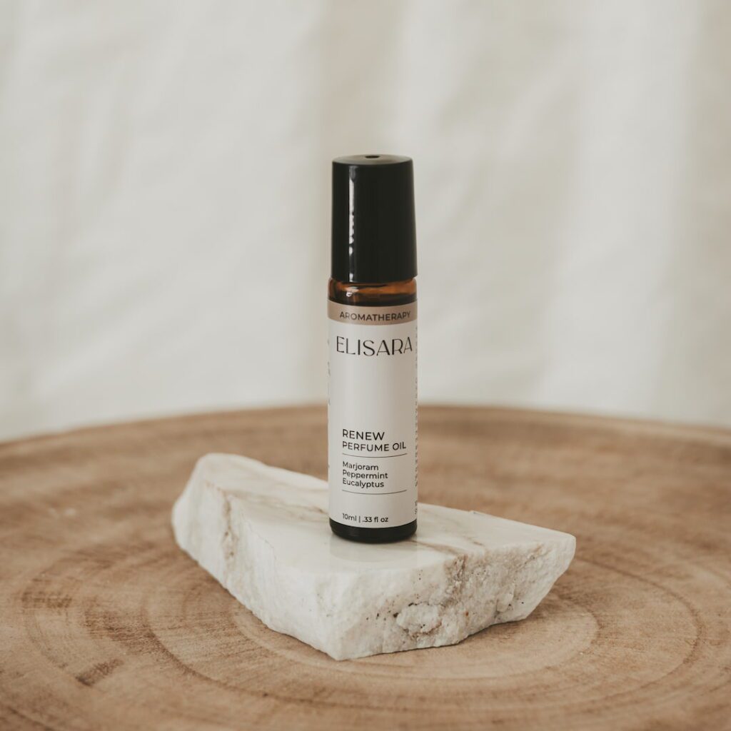 A 10ml amber roller bottle with a black lid and a white label that reads "Renew perfume oil" with the ingredients marjoram, peppermint and eucalyptus listed. The bottle is placed on a flat, white quartz stone, resting on a round timber stool. In the background, there is a white fabric curtain.