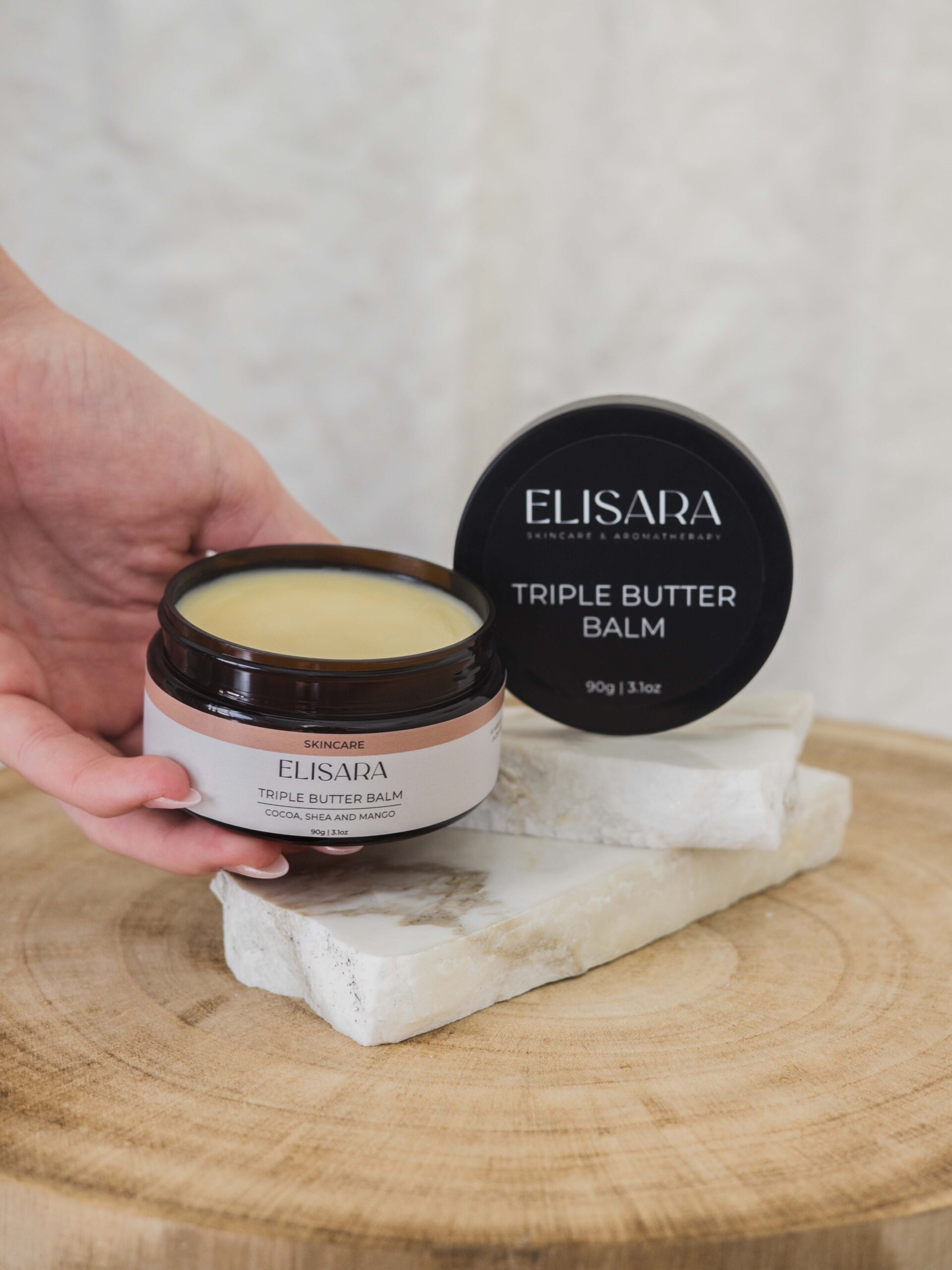 Image of a jar of triple butter balm in an amber recycled plastic tub being held by a hand. The top label is visible on the jar lid behind the hand and it all sits just about a timber stool.