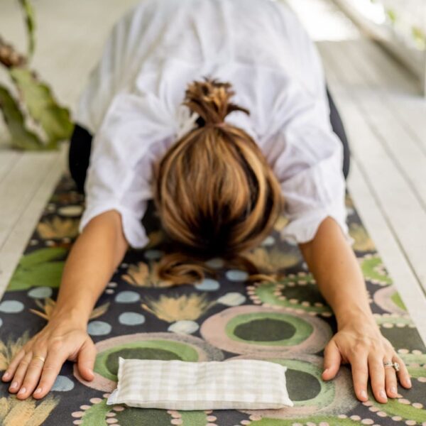 Image of a woman in downward dog pose on a yoga mat with a gingham eye pillow in between her hands which are flat of the green patterned yoga mat