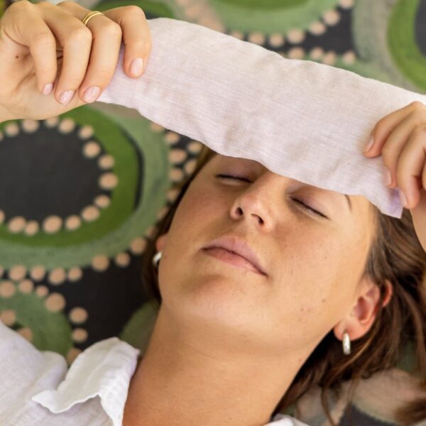 Image of a woman (face mostly) lying on a yoga mat about to apply an eye pillow in a soft pink blush colour to her eyes.