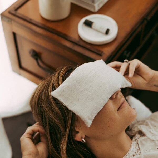 A woman lying down with her hands relaxed around her face with her eyes covered in an organic linen eye pillow. Filled with aromatherapy she looks relaxed and calm