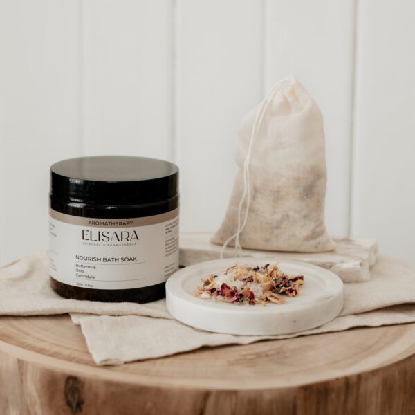 Image of a amber recycled plastic jar with black lid and a label that says Nourish bath soak with Buttermilk, oats and calendula. To the right is a reusable cotton bath tea bag and a pile of the bath salts showing herbs including rose and calendula.