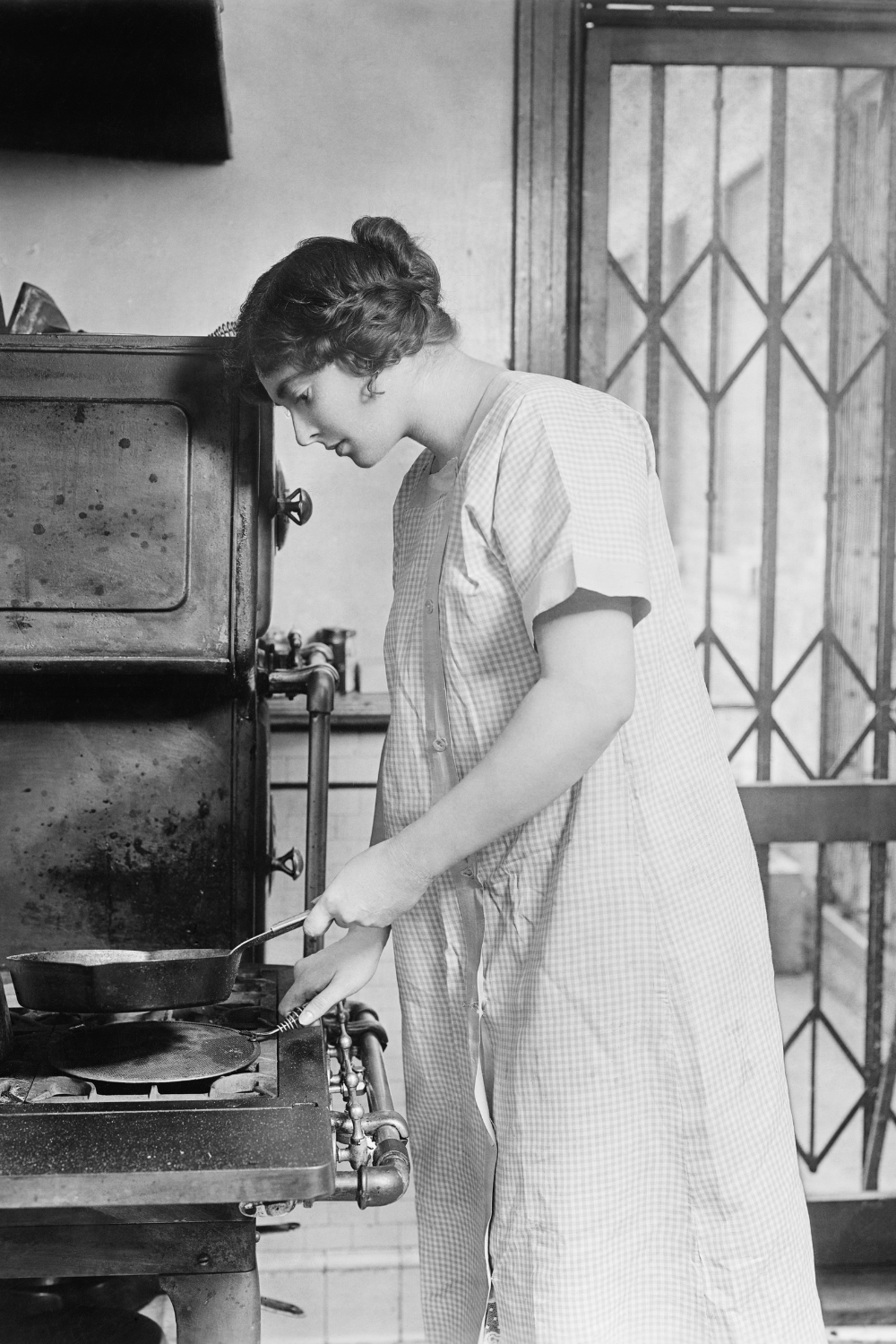 Black and white image of a woman in a older style housecoat cooking on an old fashioned cooker<br />
